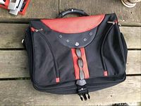 Front view of black and red laptop bag showing padded handle, zippered compartments, buckle closure and front detail with grommets.