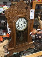 Full view of ornate gingerbread style solid wood mantle clock with carved scroll and floral details, standing on glass table surrounded by miscellaneous items.