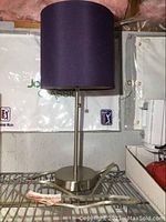 Front view of the table lamp showing the purple fabric shade and brushed metal stand and base on a wire rack.