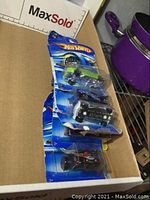 Five Hot Wheels cars stacked in box, packaging shows damage but cars sealed inside.