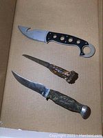 Three cutting utensils including one with a black handle and gut hook blade, one medium-sized knife, and one small knife with bone handle.