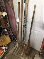 Photo showing old wooden paddle standing beside a collection of six older hockey sticks with various degrees of wear and damaged plastic ends.