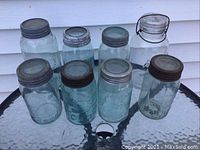 Eight vintage glass sealers displayed on a round glass table, including green glass jars with metal lids and one clear jar with a clamp lid