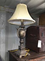 Full view of the antique style table lamp with cream fabric shade on top and urn-shaped base with square pedestal and ornate feet.