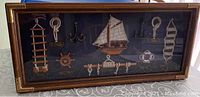 Glass framed display showing various nautical knots on rope, miniature wooden sailboat, ship wheel, anchor, lifebuoy, and rope ladders arranged on dark fabric background in wood-grain frame.