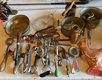 Full view of antique kitchen utensils including hand mixers, strainers, cake pans, mashers, copper measuring cups, a hen mold, and other vintage tools on a table.