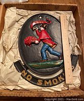 Top view of the cast iron sign in original box, showing the firefighter figure and 'NO SMOKING' text with a ruler for size reference.