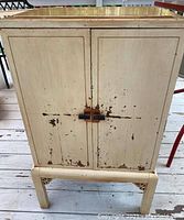 Front view of the antique painted wooden cabinet showing two doors with chipping cream paint and a wooden latch.