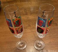 Two clear champagne flute glasses with colorful Andy Warhol Marilyn Monroe pop art images around the bowl and black signature style writing on the side, on a wooden surface.
