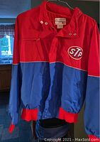 Front view of red and blue NASCAR jacket with STP logo on chest, showing color blocking, zipper, and pocket details.