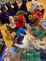 Photo showing multiple purple Princess Beanie Babies, assorted animal beanie babies including a rooster, pelican, tiger, and more, two packaged Erin Mini Beanie Babies, one packaged mini Glory Beanie Baby, and partially visible packaging.