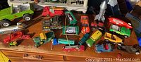 Wide view of the collection of toy vehicles including trucks, helicopters, a rocket, and a blimp on a wooden surface under sunlight.