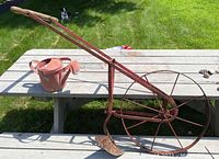 Side view of vintage farm plow showing long wooden handle, metal spoked wheel approximately 2 feet diameter, plow blade with red paint remnants, and modern metal watering can next to it on picnic table outdoors on grass.