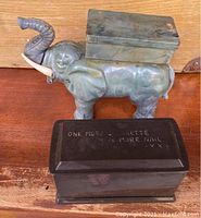 Photo of the green elephant cigarette dispenser holding a metal box on its back next to the black coffin dispenser with inscription on the lid.