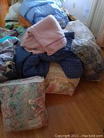 Photo capturing a pile of bedding including comforters, throw blankets, bedspreads, bed pillows and throw pillows all stacked on a wooden floor near a window.