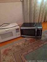 Two microwaves side by side on wooden floor: white Emerson microwave and black Darby microwave