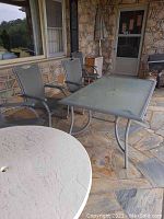 Rectangular glass top metal frame patio table with two matching metal mesh sling chairs on porch with stone floor and wall