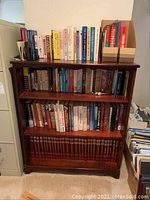 Books displayed on a wooden bookshelf with three shelves showing a variety of titles including fiction, biography, and encyclopedia volumes.