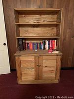 Frontal image showing the two-piece rustic wood cabinet with a closed bottom cabinet with two doors and an open bookcase with two shelves on top. Some books and items are placed on the shelves but not included in the lot.