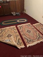 Photo showing three rectangular pink Arak rugs arranged on red carpet with fringe visible, and two oval braided rugs placed above them.