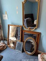 Mirrors grouped on floor against blue wall, varying gold-tone ornate frames in square, rectangular and oval shapes; also smaller framed mirrors included