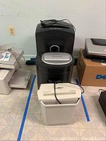 Three paper shredders seen from side and top angles, showing different models and colors, cords attached and front access handles.