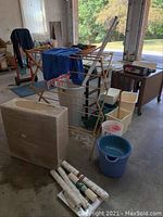 Photo showing a wooden laundry drying rack with clothing hanging on it, buckets of various sizes and colors, trash cans, and a cardboard box, along with three rolls of material on the ground.