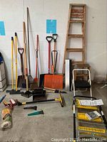 Entire lot of yard tools, ladder, stepstool, shovels, rakes, brooms, squeegees against a white wall