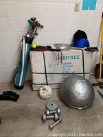 Multiple golf clubs in a blue golf bag, two helmets, badminton and racquetball rackets on a box labeled Orbitrek, with several hand weights and a silver exercise ball on the floor.