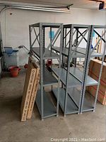 Three grey heavy-gauge steel shelves assembled side by side, showing vertical supports with bolt holes and cross braces on the back. Some surface rust visible on the assembled shelves.