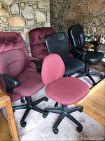 Photo showing four office chairs of different styles and colors including red fabric, black mesh, burgundy cushioned, and black leather-like cushioned chairs against a stone wall background.