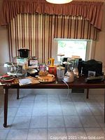 Photo showing multiple small kitchen appliances and accessories arranged on a wooden dining table.