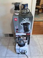 Photo showing stack of boxes with illy X8 espresso machine, Braun Tassimo coffee maker and Capresso H2O Plus electric water kettle on tiled floor.