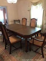 Full view of wooden dining room table with six padded chairs around it in a room with carpet and window curtains.