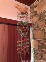 Pair of handmade Hawaiian wind chimes hanging indoors near a stone fireplace and wooden wall, showing full length and design details including wooden top, green leafy heart shape, colorful parrots, and bamboo tubes.