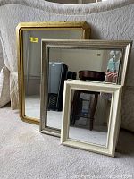 Front view of three mirrors of different sizes and colors leaning against a sofa: gold, silver, and cream wood frames.