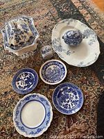 Full group of eight blue and white china pieces including plates, covered dish, small cup, and egg-shaped covered piece, displayed on a patterned carpet.