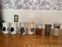 Seven handcrafted ceramic teacups of different glaze patterns and colors set on wooden floor against wall.