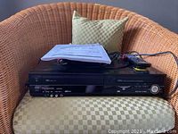 Front view of Panasonic DMR-EZ485V DVD recorder with VHS player showing front panel including disc tray, VHS slot, and AV ports. Owner's manual and remote placed on top.