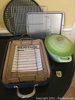 Photo of assorted cookware showing a Heritage Rock roasting pan with metal rack, two metal baking trays, a round perforated tray, and a green ceramic Dutch oven with lid on a round black table.