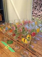 Front view showing multiple hand painted white wine glasses arranged in rows with floral designs and some green stems visible, placed on wooden surface near glass door.