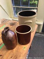 Three earthenware pots on wooden table near window: brown glazed jug, beige stoneware crock with crown marking, smaller brown glazed crock.