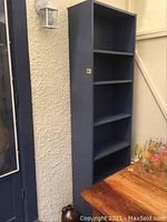 Front and angled view of the empty Wedgwood blue 6-shelf bookshelf against an exterior textured wall near a wooden table with glassware, showing shelf spacing, paint wear, and overall structure.