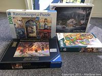 Four boxed puzzles on carpet: Ravensburger 1500-piece, Ravensburger Special Edition 1000-piece, indigenous art puzzle by Sue Coccia 1000-piece, and a winter cabin scene puzzle with 1000 pieces.