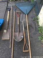 Photo showing six garden tools including shovels, forks, rakes with wooden handles and metal heads placed on the ground.
