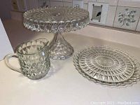 Three glass items: cake stand, plate, and small pitcher displayed on a countertop.