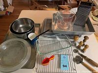 Overview of various kitchen and baking pans on table including metal bowls, pie pan, baking sheets, glass measuring cup, glass baking dish, wire rack, and wooden-handled tools.