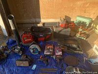 Overview on blue tarp showing two belt sanders, circular saw, reciprocating saw, chainsaw, power drill, leaf blower, drill press, files and tool bag