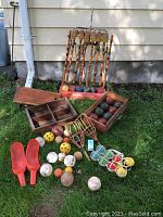 Complete view of croquet mallets, croquet balls in the red 'Lawnplay' stand, wooden bocce ball box with bocce balls, various balls, red ball scoops, and batting catchers arranged on grass.