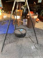 Cast iron Dutch oven hanging from metal tripod with chain, shown outdoors on pavement near garage.